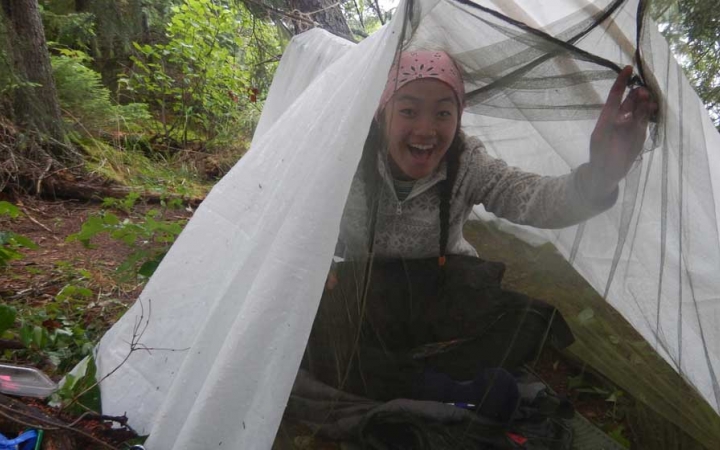 a person smiles from under a tarp shelter amongst trees on an outward bound course